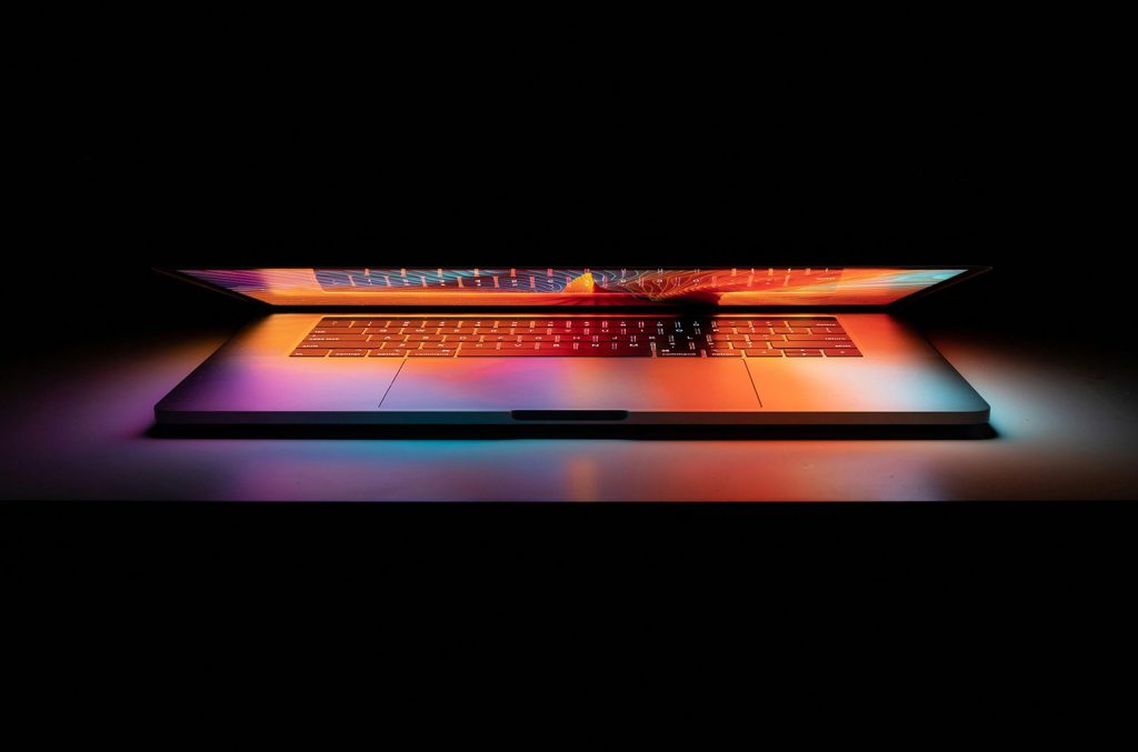 Image of laptop partially open, with a colorful glow around the keyboard; on a black background
